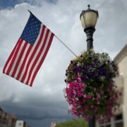Photo of the US flag flying near a hanging flower basket.